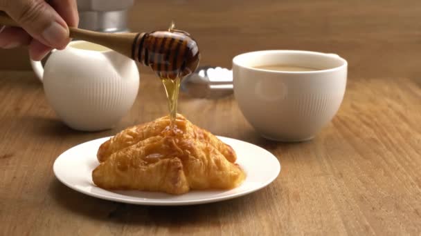 用蜂蜜调酒师将蜂蜜倒入白色陶瓷盘中美味的自制牛角面包 并在木桌上倒入一杯热牛奶咖啡 — 图库视频影像