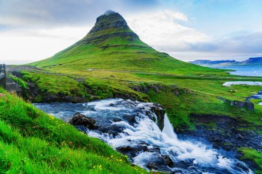 Kirkjufellsfoss Şelaleleri ve Kirkjufell Dağı 'nın doğal manzarası. Yaz mevsiminde yeşil çimen ve sarı çiçeklerin olduğu şafak vakti İzlanda..