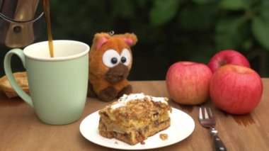 Pouring hot black coffee into a green ceramic cup with delicious homemade fruit cake in white ceramic dish and red rip apple on wooden table.