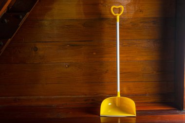Long handle plastic yellow dustpan leaning against wooden wall on a wooden floor in a room.