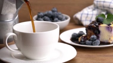 Closeup of pouring hot black coffee into white ceramic cup on white saucer with a slice of blueberries yoghurt cake pouring with chocolate syrup and blueberries fruit on wooden table.