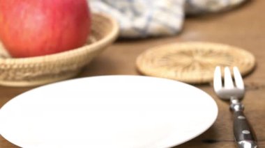 Putting a portion of homemade walnut fruitcake in white ceramic dish and placing a glass of orange juice on bamboo mat with ripe red apple in bamboo basket on wooden table.
