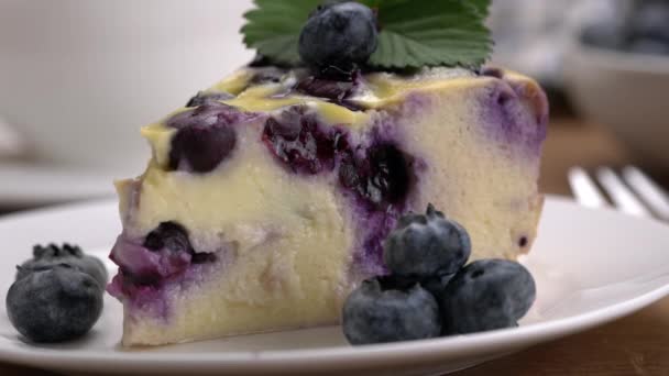 在白色陶瓷盘中 将巧克力糖浆倒入蓝莓酸奶蛋糕片 并将蓝莓水果放在木制桌子上的极端特写镜头 — 图库视频影像