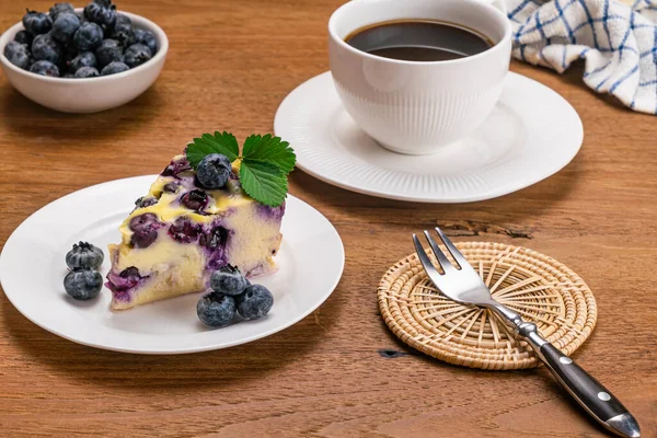 Blueberry yoghurt cake and black coffee in coffee time. Slice of blueberry yogurt cake in white ceramic dish with a cup of black coffee on white saucer and blueberry fruit in a ceramic cup on wooden table.