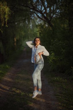 Seksi, şehvetli esmer, yaz ormanlarında gün batımında beyaz bluz ve kotla yürüyor..