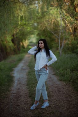 Seksi, şehvetli esmer, yaz ormanlarında gün batımında beyaz bluz ve kotla yürüyor..