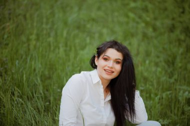 Seksi esmer, beyaz bluz ve kotla bir yaz parkında çimlerin üzerinde oturuyor..