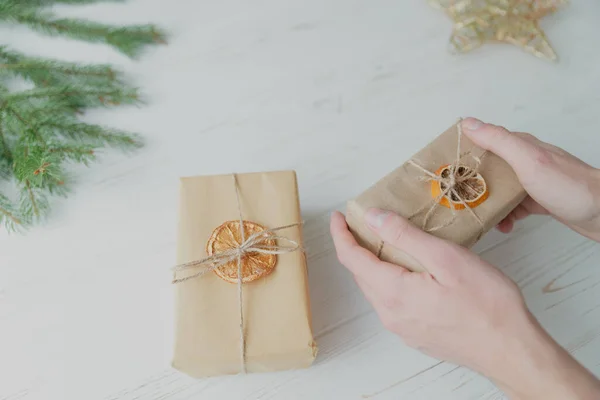A man wraps a Christmas present in craft paper on a white wooden background.