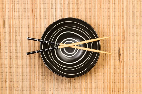 Chopsticks on a black bowl and on a bamboo table mat in a top view