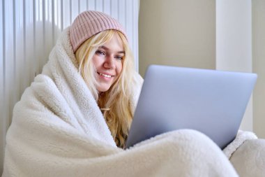 Soğuk kış mevsiminde donan ergen, battaniye şapkasıyla ısınıyor, dizüstü bilgisayara bakıyor.