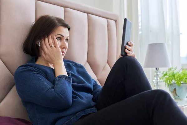 Traurige Frau mittleren Alters mit Kopfhörern, die in den Smartphone-Bildschirm blickt. — Stockfoto