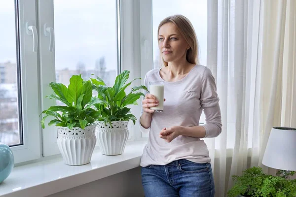 Frau mittleren Alters mit einem Glas Milch, zu Hause am Fenster — Stockfoto