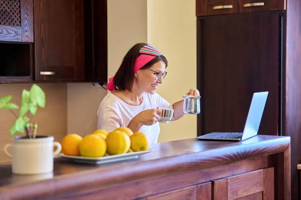 Kvinna i köket med en kopp kaffe tittar in i den bärbara skärmen — Stockfoto