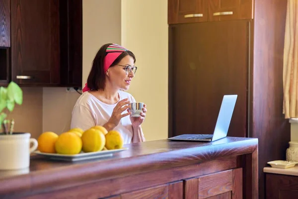 Mujer en la cocina con una taza de café mirando en la pantalla del ordenador portátil — Foto de Stock