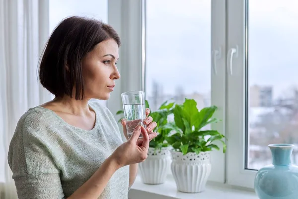 Mujer de mediana edad con píldora y vaso de agua en las manos, en casa en invierno — Foto de Stock