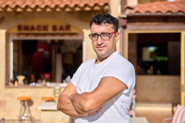 Kendinden emin orta yaşlı, çapraz kollu gözlük takan bir adamın portresi.