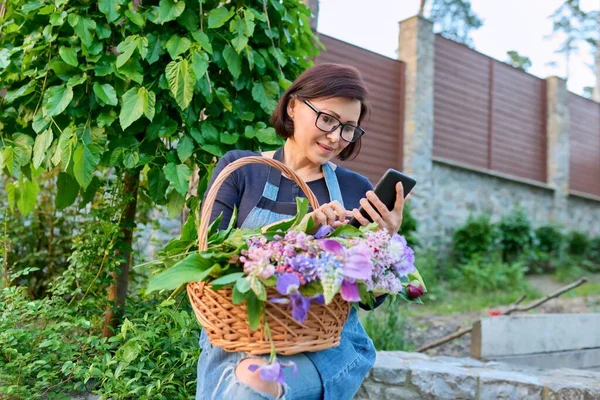 Μεσήλικη γυναίκα στον κήπο με καλάθι φρέσκα ανοιξιάτικα λουλούδια κομμένα με smartphone. — Φωτογραφία Αρχείου
