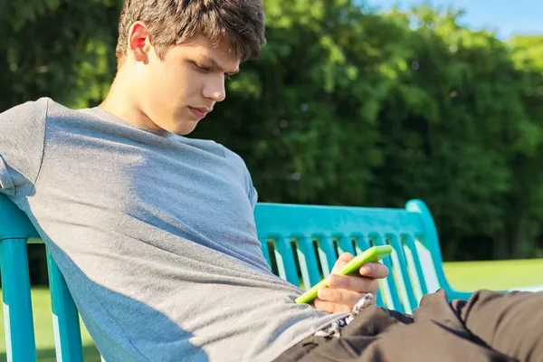Chico serio adolescente usando smartphone, descansando, sentado en el banco en el parque — Foto de Stock