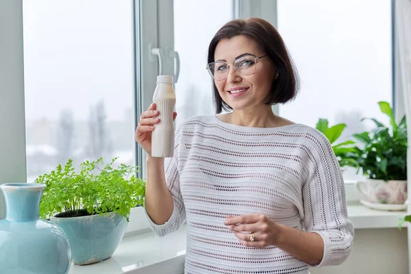 Frau mittleren Alters trinkt Milchgetränk, flüssigen Joghurt in der Flasche, zu Hause am Fenster — Stockfoto