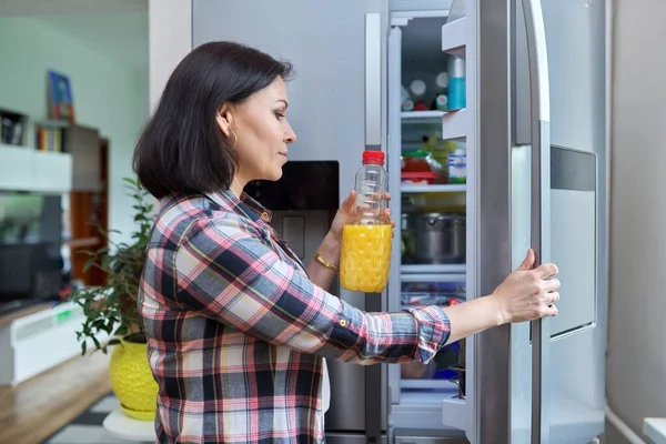 A woman opens the refrigerator at home in the kitchen, holding a bottle of orange juice.