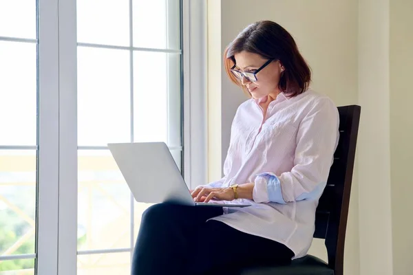 Donna d'affari matura in occhiali con computer portatile nelle sue mani vicino alla finestra — Foto Stock