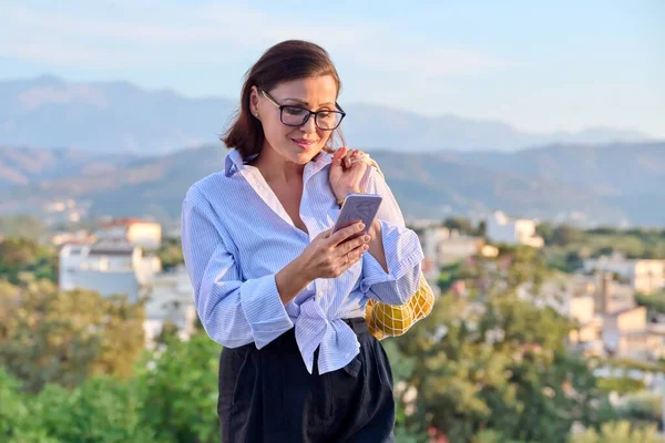 Retrato de mulher de meia-idade com smartphone e grade de tendências de laranjas ao ar livre — Fotografia de Stock