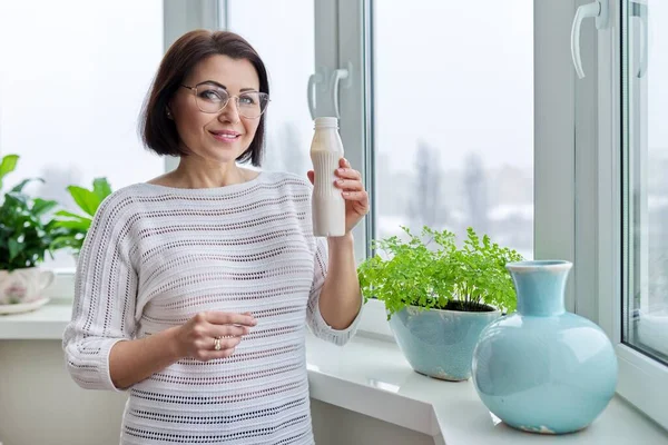 Frau mittleren Alters trinkt Milchgetränk, flüssigen Joghurt in der Flasche, zu Hause am Fenster — Stockfoto