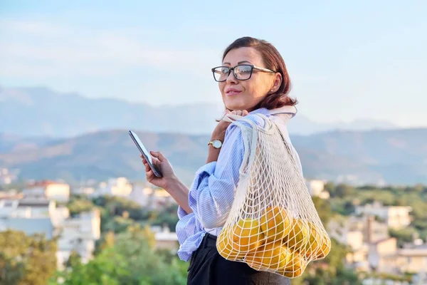 스마트폰을 들고 있는 중년 여성의 모습 과 밖에서 오렌지를 포장하고 있는 모습 — 스톡 사진