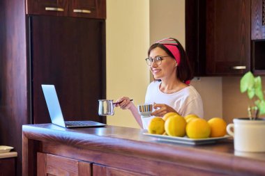 Gülümseyen kadın, sabah kahvesiyle evdeki mutfakta Türk ve dizüstü bilgisayarla rahatlıyor.