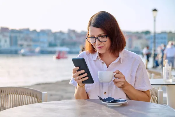 Жінка середнього віку відпочиває у відкритому кафе з чашкою кави за допомогою смартфона — стокове фото