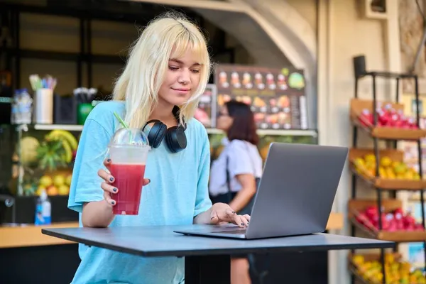 Teenage girl in fruit outdoor fruit juice bar πίνοντας φρέσκο χυμό βιταμίνης, χρησιμοποιώντας φορητό υπολογιστή — Φωτογραφία Αρχείου