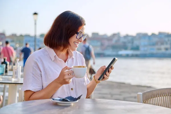 Mulher de meia-idade descansando no café ao ar livre com uma xícara de café usando um smartphone — Fotografia de Stock