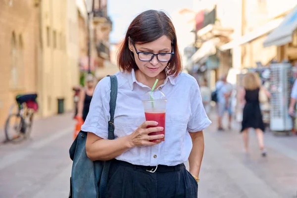 Mulher de meia-idade com copo de suco fresco nas mãos andando pela cidade — Fotografia de Stock