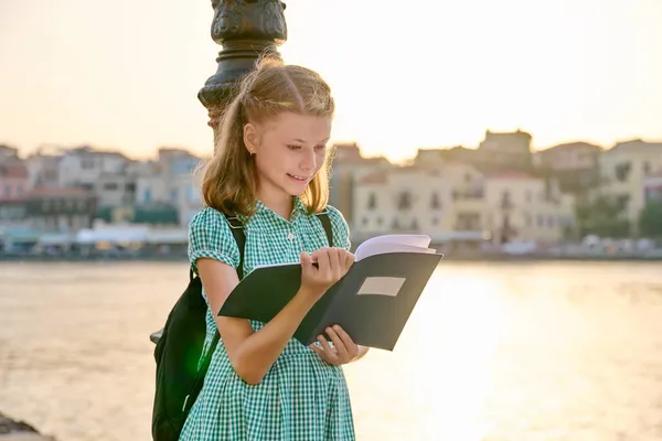 Niña estudiante joven con mochila lectura cuaderno de la escuela, al aire libre — Foto de Stock