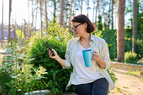 Μεσήλικη γυναίκα που ξεκουράζεται στον κήπο της άνοιξης με φλιτζάνι τσάι και smartphone στα χέρια — Φωτογραφία Αρχείου