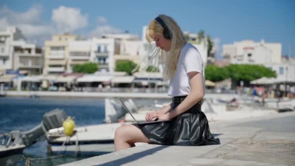 Mujer adolescente en auriculares con portátil sentado cerca del agua, puerto de mar con barcos — Vídeos de Stock