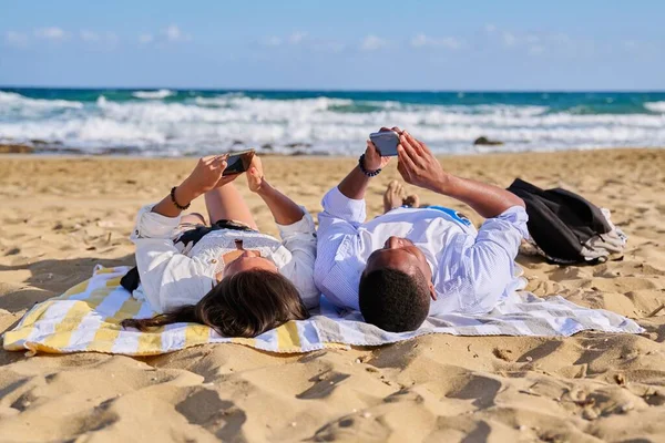 Mladý multikulturní pár ležící na pláži pomocí chytrých telefonů — Stock fotografie