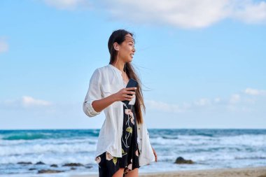 Kulaklıklı, akıllı telefonlu, plajda müzik dinleyen genç bir kadın.