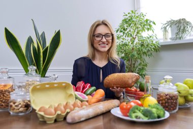 Elinde ekmek, masada yemek olan bir kadın kameraya bakıyor.