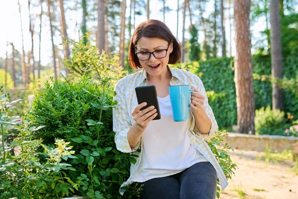 Μεσήλικη γυναίκα που ξεκουράζεται στον κήπο της άνοιξης με φλιτζάνι τσάι και smartphone στα χέρια — Φωτογραφία Αρχείου