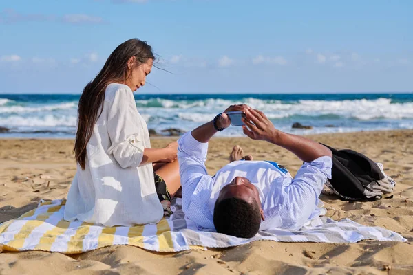 Jong multicultureel paar liggend op het strand met behulp van smartphones — Stockfoto