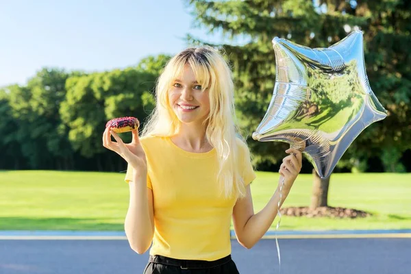 Portret van mooie tiener blond met cake donut en zilveren ballon in het park — Stockfoto