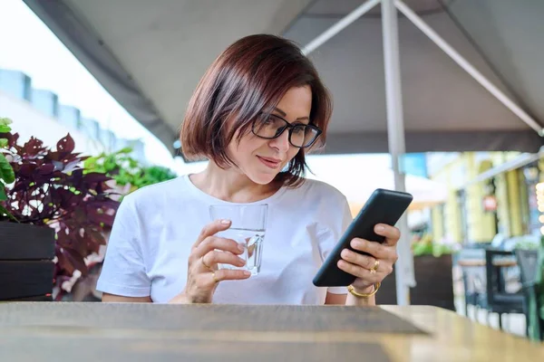 40s female sitting in an outdoor cafe with glass of water using smartphone — Stock Photo, Image