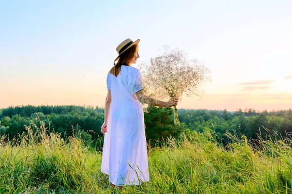 美丽的女人，头戴白色礼帽，花束缀在草地上，日落时分 — 图库照片