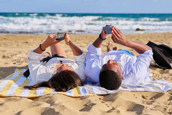 Fiatal multikulturális pár fekszik a tengerparton okostelefonnal — Stock Fotó