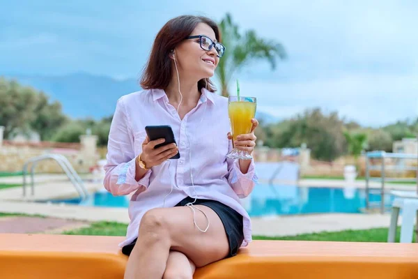 Женщина средних лет отдыхает у бассейна, используя наушники и смартфон — стоковое фото