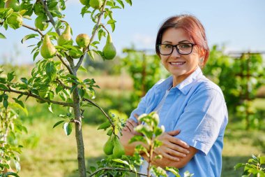 Armut ağacının yanında, meyve bahçesinde çapraz kollu, kendinden emin orta yaşlı bir kadının portresi.