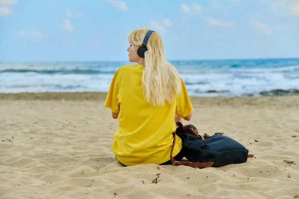 Adolescente femenina en auriculares con mochila sentada en la playa, vista trasera, espacio para copiar — Foto de Stock