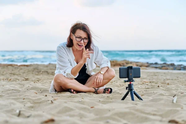 Μεσήλικη γυναίκα κάθεται στην παραλία με smartphone χρησιμοποιώντας βιντεοκλήση — Φωτογραφία Αρχείου