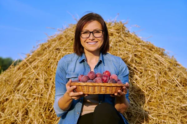 Summer rural woman portrait with basket of plums, dry hay sky background — Stock Photo, Image